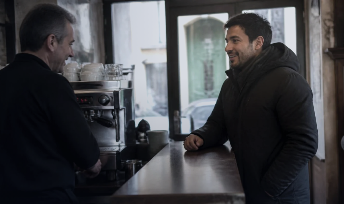 Homem jovem apoiado no balcão de um bar italiano conversa com o barista, com a máquina de café e a porta de vidro da rua ao fundo