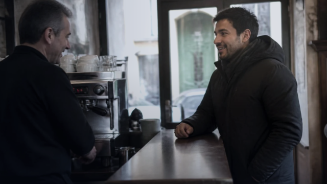Homem jovem apoiado no balcão de um bar italiano conversa com o barista, com a máquina de café e a porta de vidro da rua ao fundo
