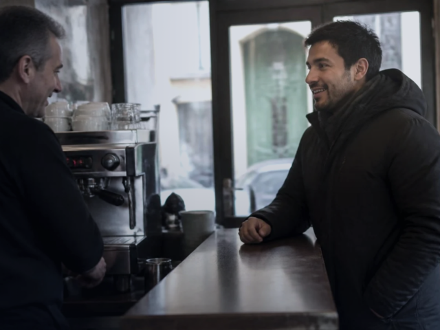 Homem jovem apoiado no balcão de um bar italiano conversa com o barista, com a máquina de café e a porta de vidro da rua ao fundo